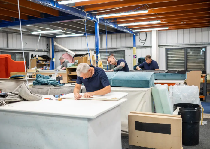 Workers in an upholstery workshop measuring and assembling materials at large tables with tools, fabric rolls, and cushions around them.