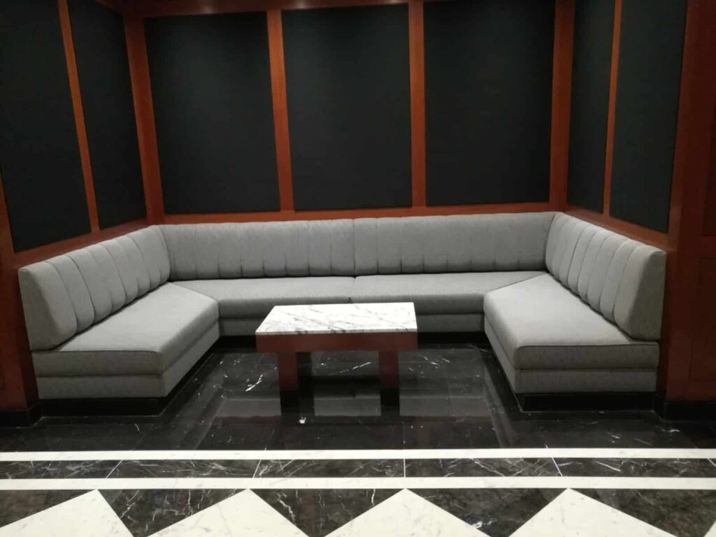 A large, custom ushaped banquette upholstered in grey fabric with channel tufting, installed in a lounge room with dark wood and black acoustic wall panels.