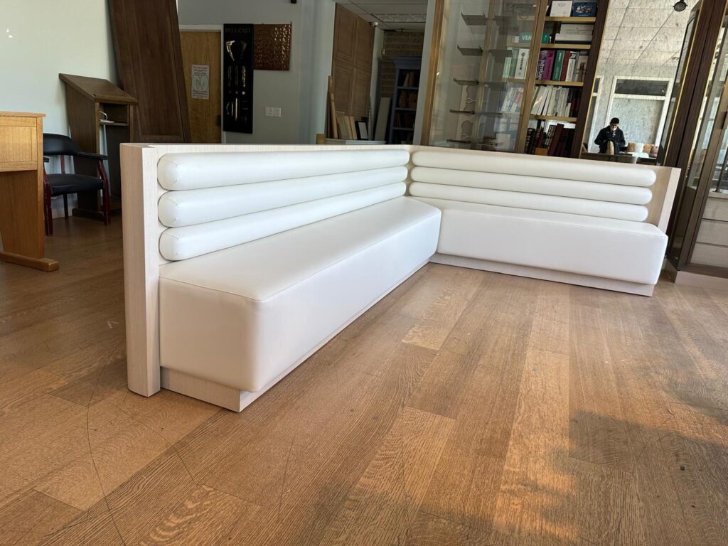 Custom lshaped corner banquette sofa upholstered in white leather, featuring horizontal channel stitching on the backrest, placed on a wideplank wood floor in an interior setting.