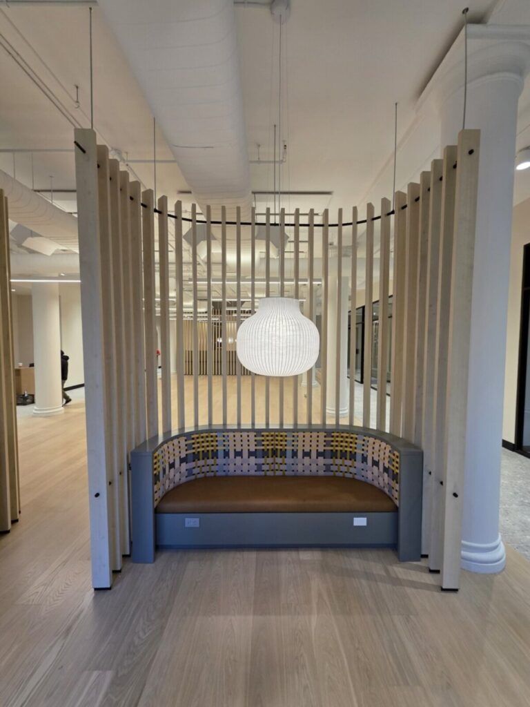 A custom curved banquette with grey upholstery, backed by a slatted wood wall panel divider, situated in a modern open office space with a white globe pendant light overhead.