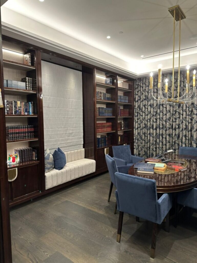 A custom white upholstered banquette seat built directly into a wall of dark wood bookcases in an elegant library, featuring a round table, blue velvet dining chairs, and a modern chandelier.