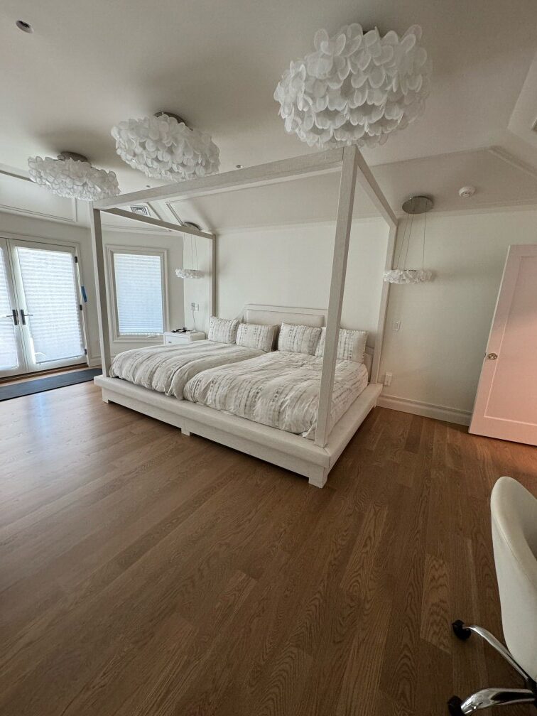 Completed white canopy bed with padded headboard installed in a finished bedroom featuring wood flooring and decorative ceiling fixtures.