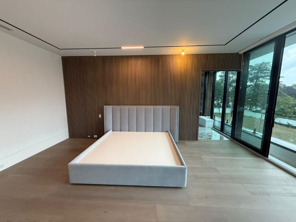 Completed modern bed and headboard installed against a wood accent wall in a minimalist bedroom with floor‑to‑ceiling windows.