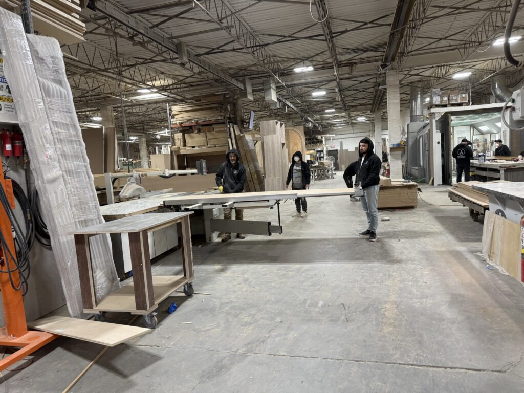 Multiple workers in the shop moving large wooden panels and components during active bed and headboard construction.