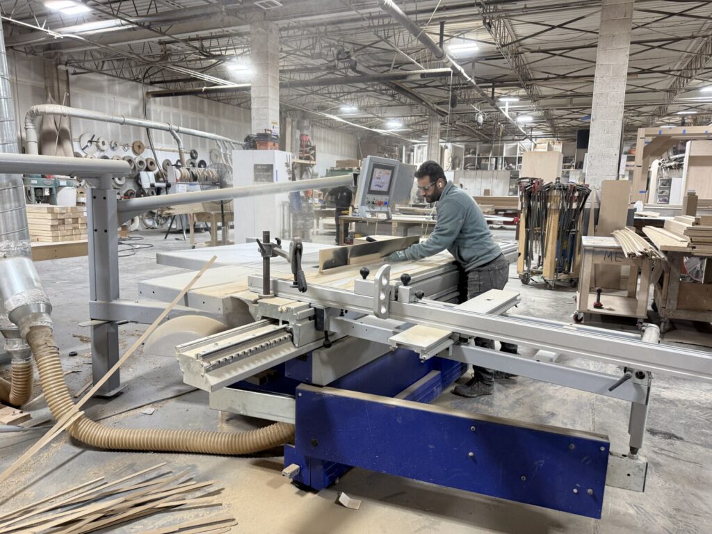 Technician operating a cnc router in a spacious workshop with wood pieces and dust‑collection hoses during active bed fabrication.
