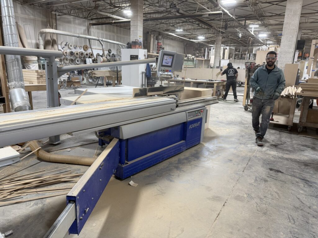 A woodworking shop with an omga industrial cutting machine processing wooden planks, surrounded by sawdust, stacked lumber, and workshop equipment while two workers walk through the fabrication area.