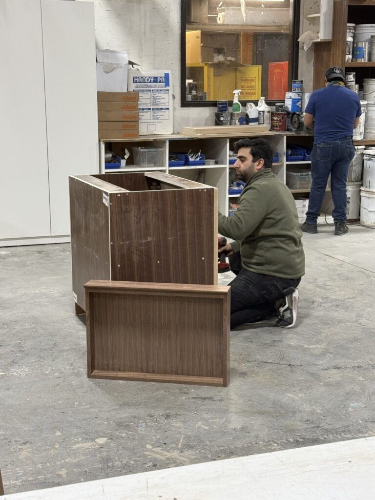 Worker kneeling to assemble a partially built wooden bed frame while another technician works at a bench in a stocked workshop.