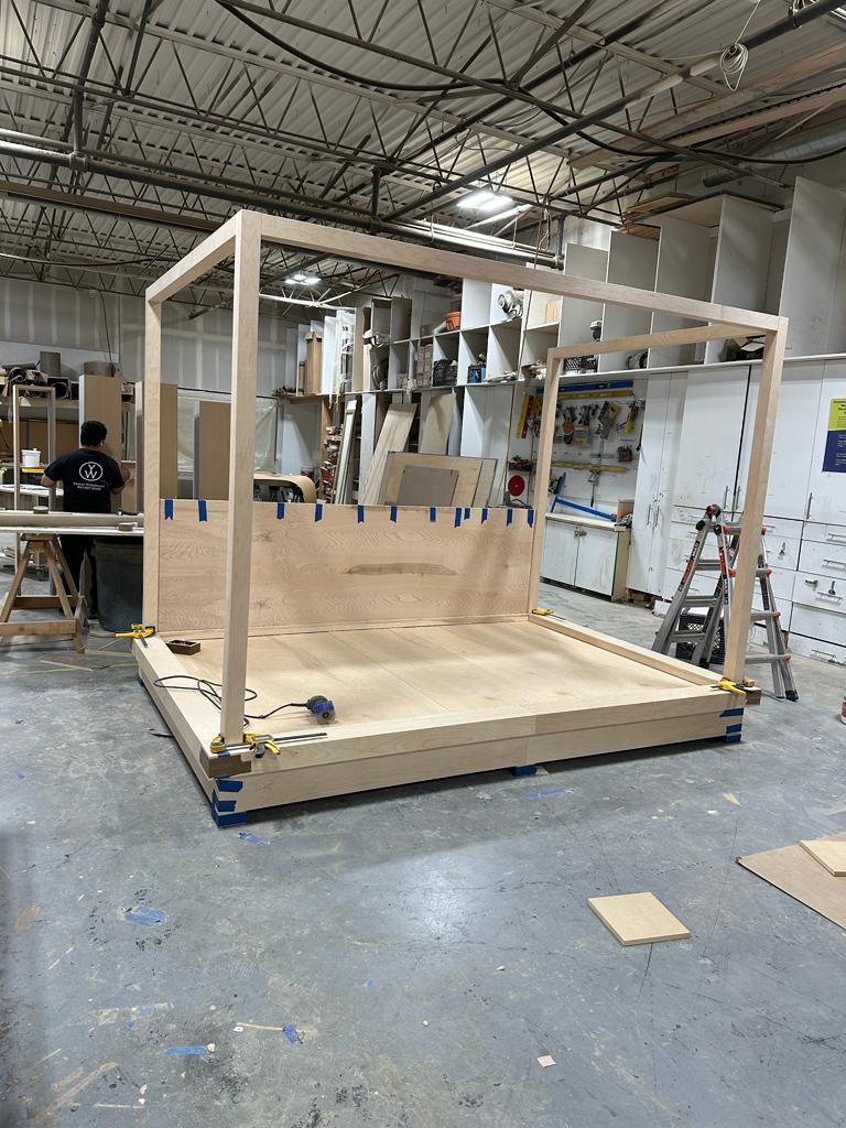 Large wooden bed frame with canopy structure being assembled in a fabrication shop, tools and materials surrounding the workspace.