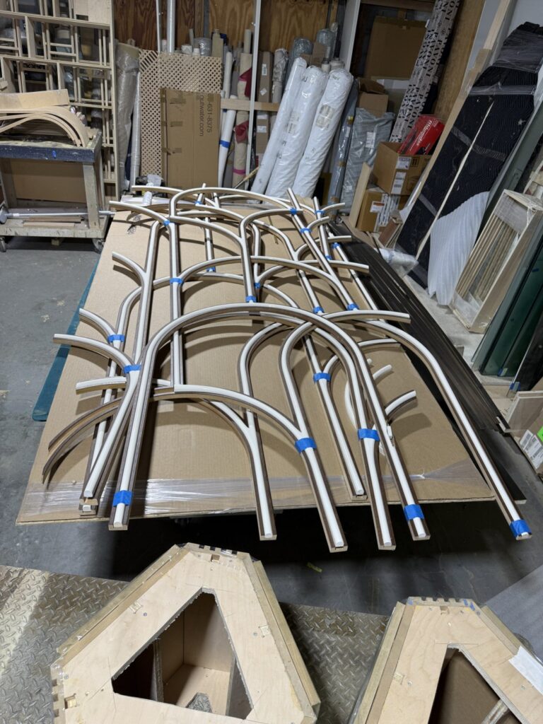 Stack of curved tubular metal parts arranged on a workbench in a fabrication shop with tools and materials in the background.