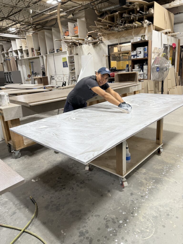 Technician applying material onto a large rectangular panel in a busy workshop with tools and supplies.