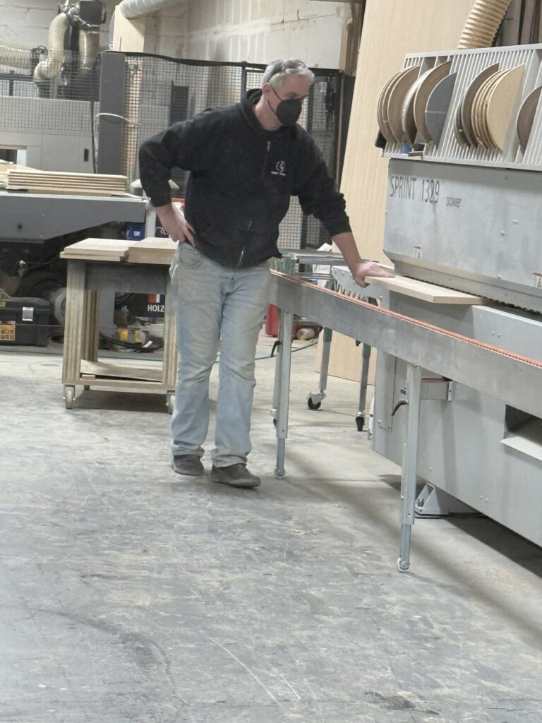 Technician standing beside a long wood piece positioned on a large workshop machine used for sanding or edge processing.