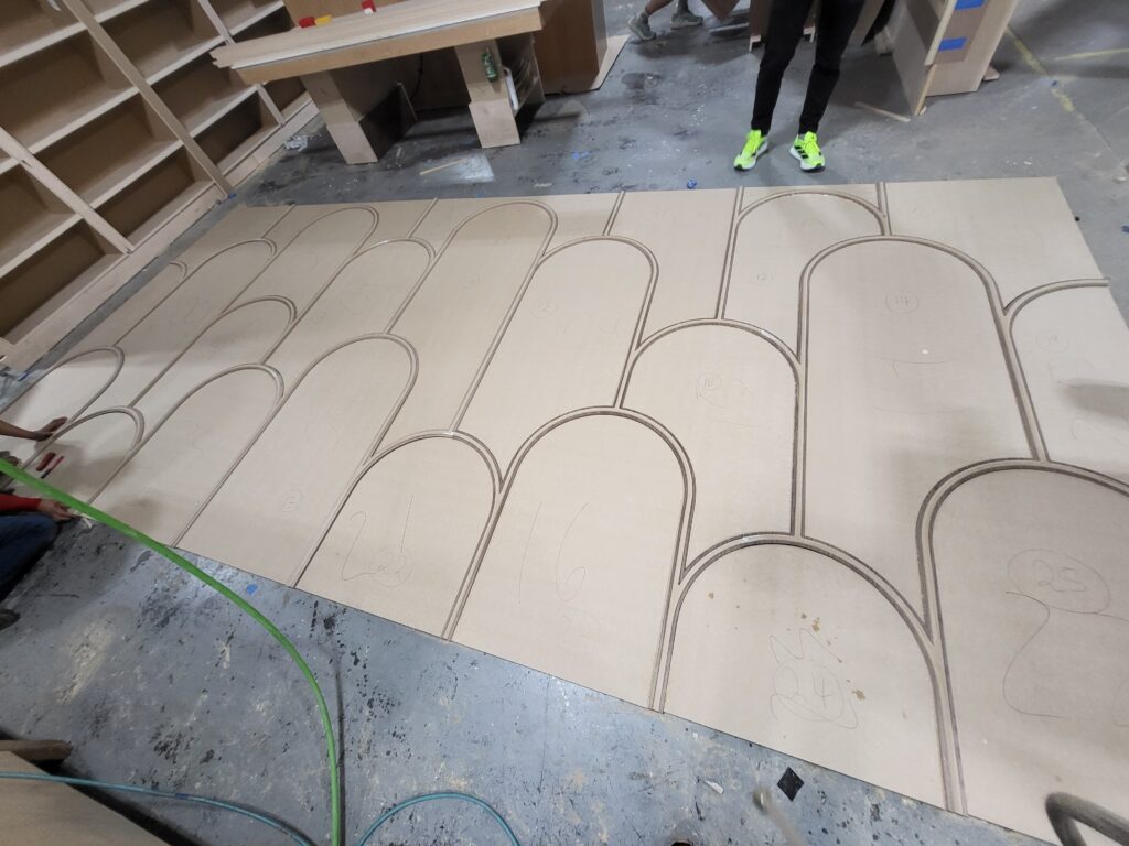 Large mdf sheet on workshop floor with arched cnc‑cut patterns; technician’s legs visible behind the panel.
