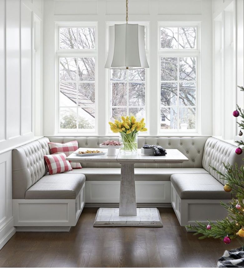 A bright u‑shaped kitchen‑nook banquette with tufted white upholstery, a pedestal dining table, striped accent pillows, and a vase of yellow flowers.